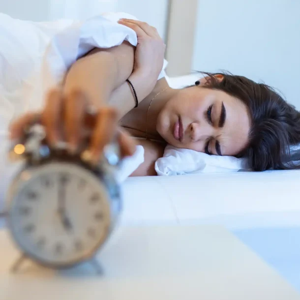 Woman lying in bed reaching for an alarm clock, appearing tired and sleep-deprived, symbolizing the impact of insomnia and poor sleep on brain and health - Save your Brain Wellness