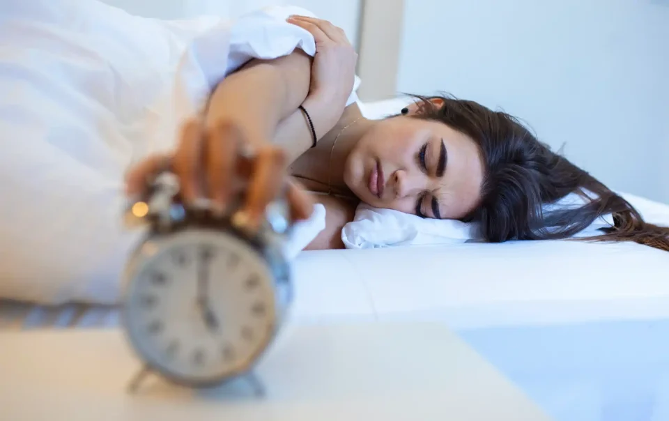 Woman lying in bed reaching for an alarm clock, appearing tired and sleep-deprived, symbolizing the impact of insomnia and poor sleep on brain and health - Save your Brain Wellness